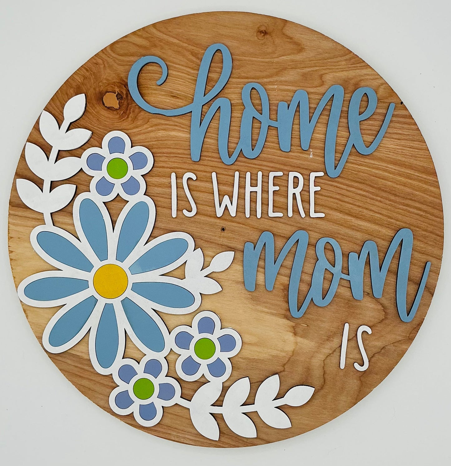 Wooden circular sign with floral designs and text 'home is where mom is' on a white background