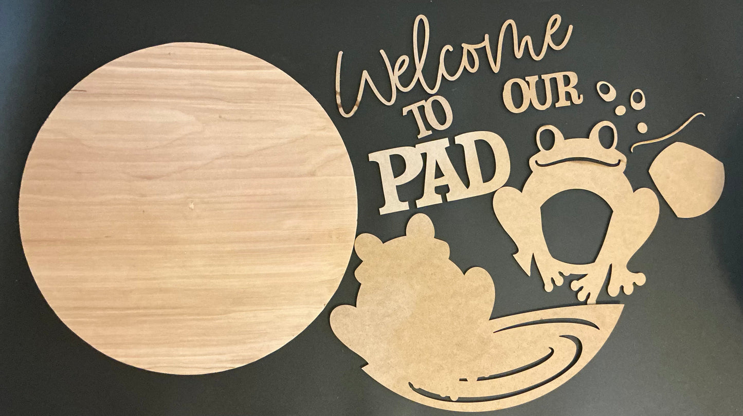 Wooden cutout with 'Welcome to Our Pad' text and frog design on a dark background
