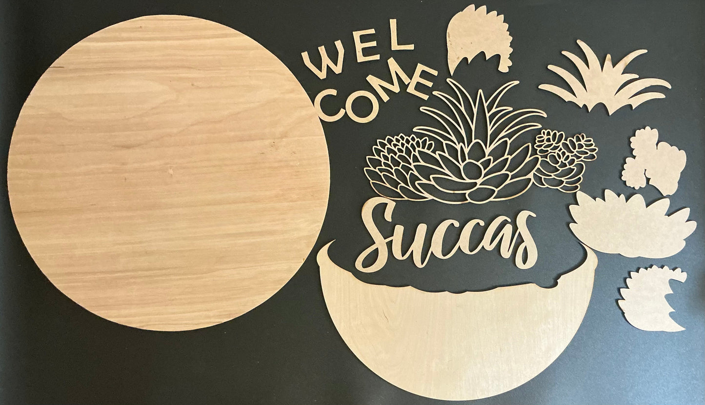 Wooden cutout with 'Welcome Succas' text and succulent designs on a black background