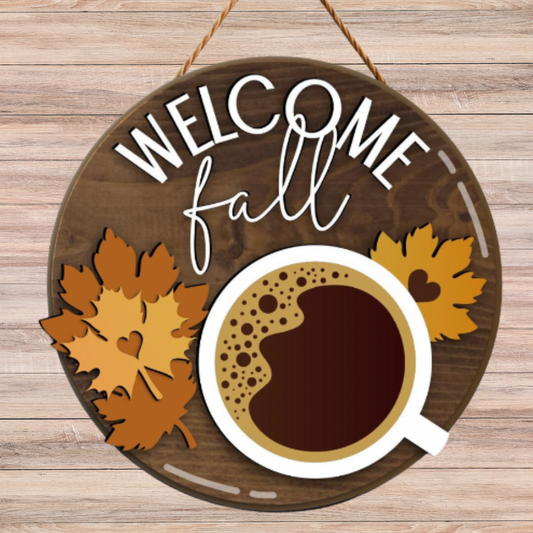 Round wooden hanging sign with the words “WELCOME fall” in white lettering, decorated with autumn leaves and a coffee cup illustration, shown against a light wood plank background.