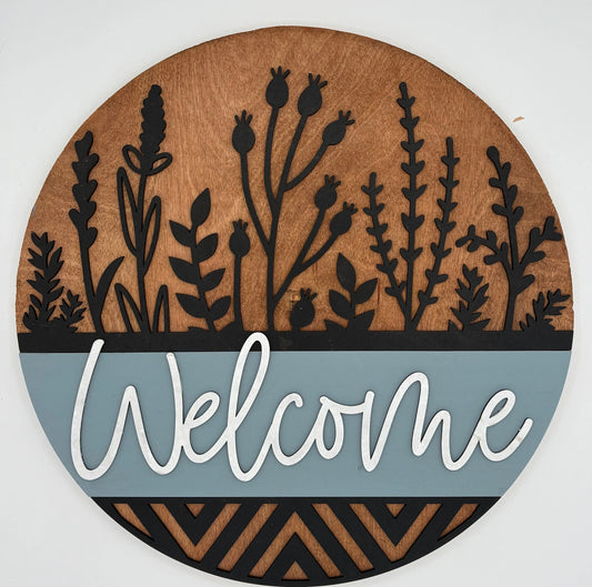 Round wooden sign with black plant silhouettes, 'Welcome' text, and geometric patterns on a white background