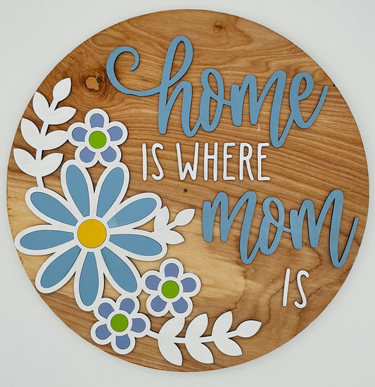 Wooden circular sign with floral designs and text 'home is where mom is' on a white background