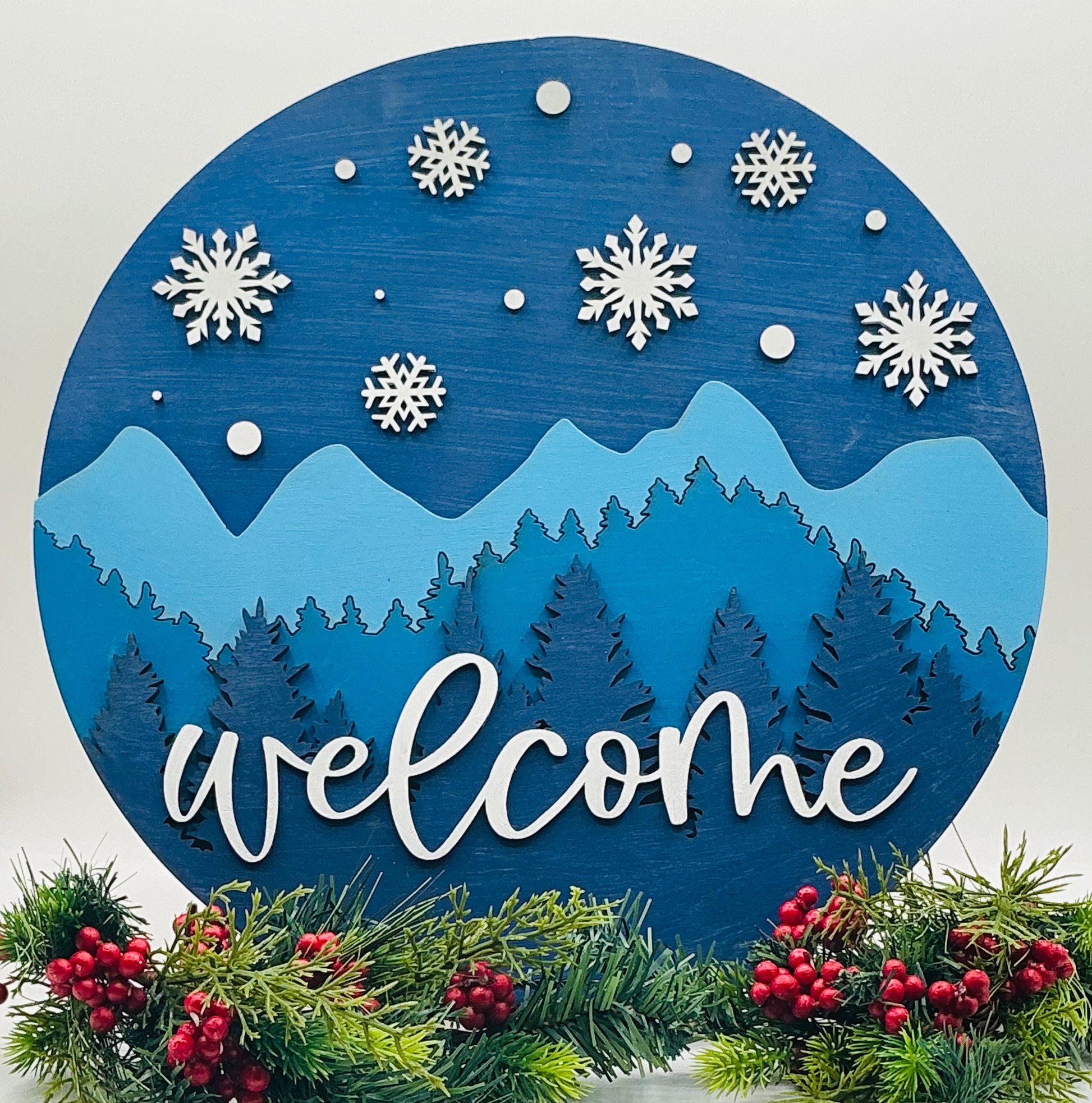 A circular decorative 'Welcome' sign featuring snowflakes and a mountain range in blue and white colors, surrounded by a border of red berries and green foliage.