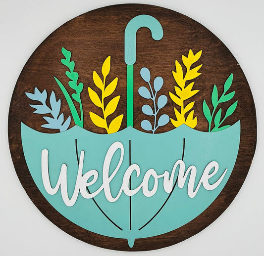 Decorative sign with 'Welcome' text inside a stylized umbrella with leaf designs on a wooden background.