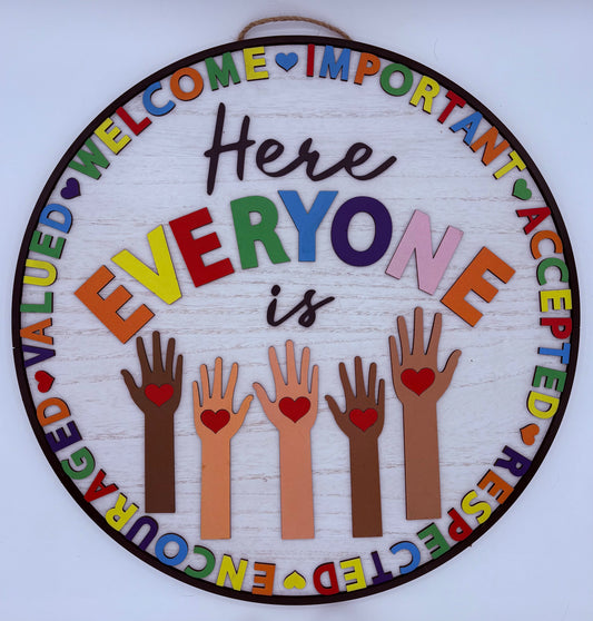 Round wooden sign with colorful text saying "Here everyone is" with descriptive words around with hands holding hearts on a white background