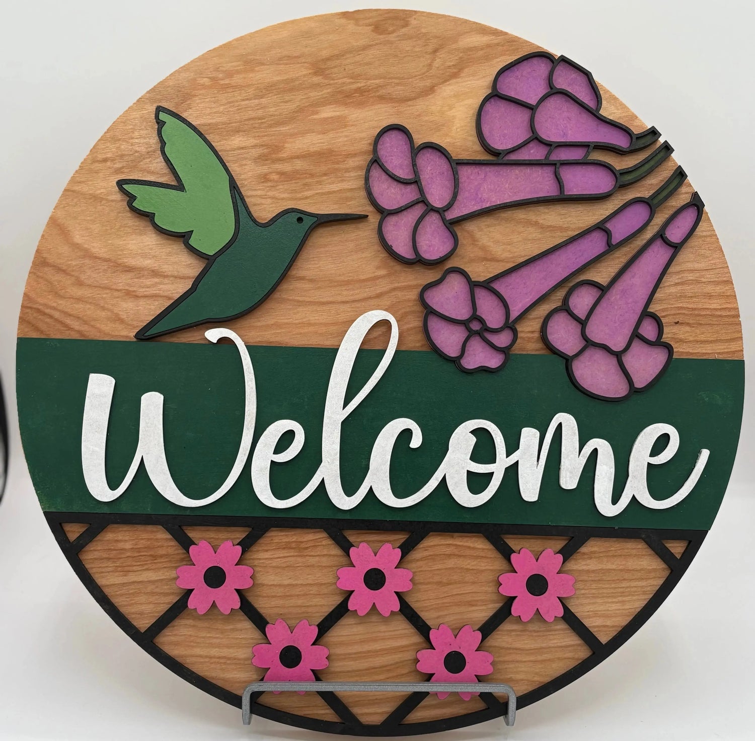 Round wooden sign with a green hummingbird, pink flowers, and the word 'Welcome' on a white background.