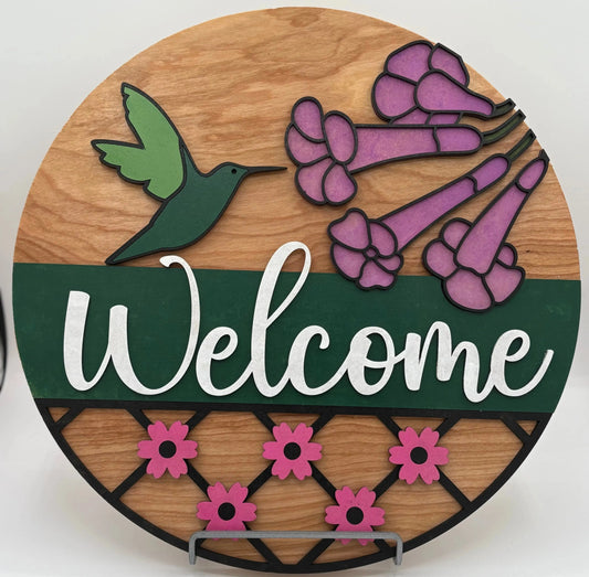 Round wooden sign with a green hummingbird, pink flowers, and the word 'Welcome' on a white background.